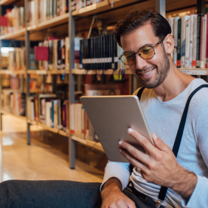 An adult man reading a tablet with bookshelves behind him. 