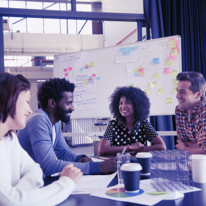 Image of a group of people sitting around a table with a white board, notes, and coffee