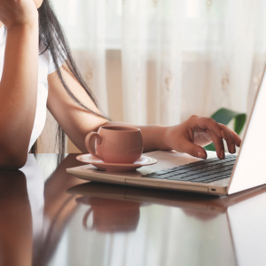 An image of a female with hands on a laptop and a mug
